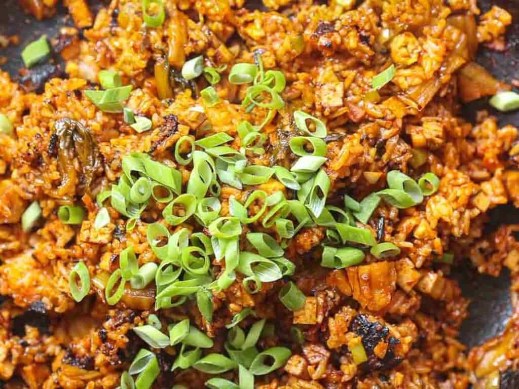 A close-up of spicy vegan kimchi fried rice mixed with kimchi, garnished with chopped green onions. The rice is vibrant orange-red, indicating rich spices, and slightly charred for added texture.
